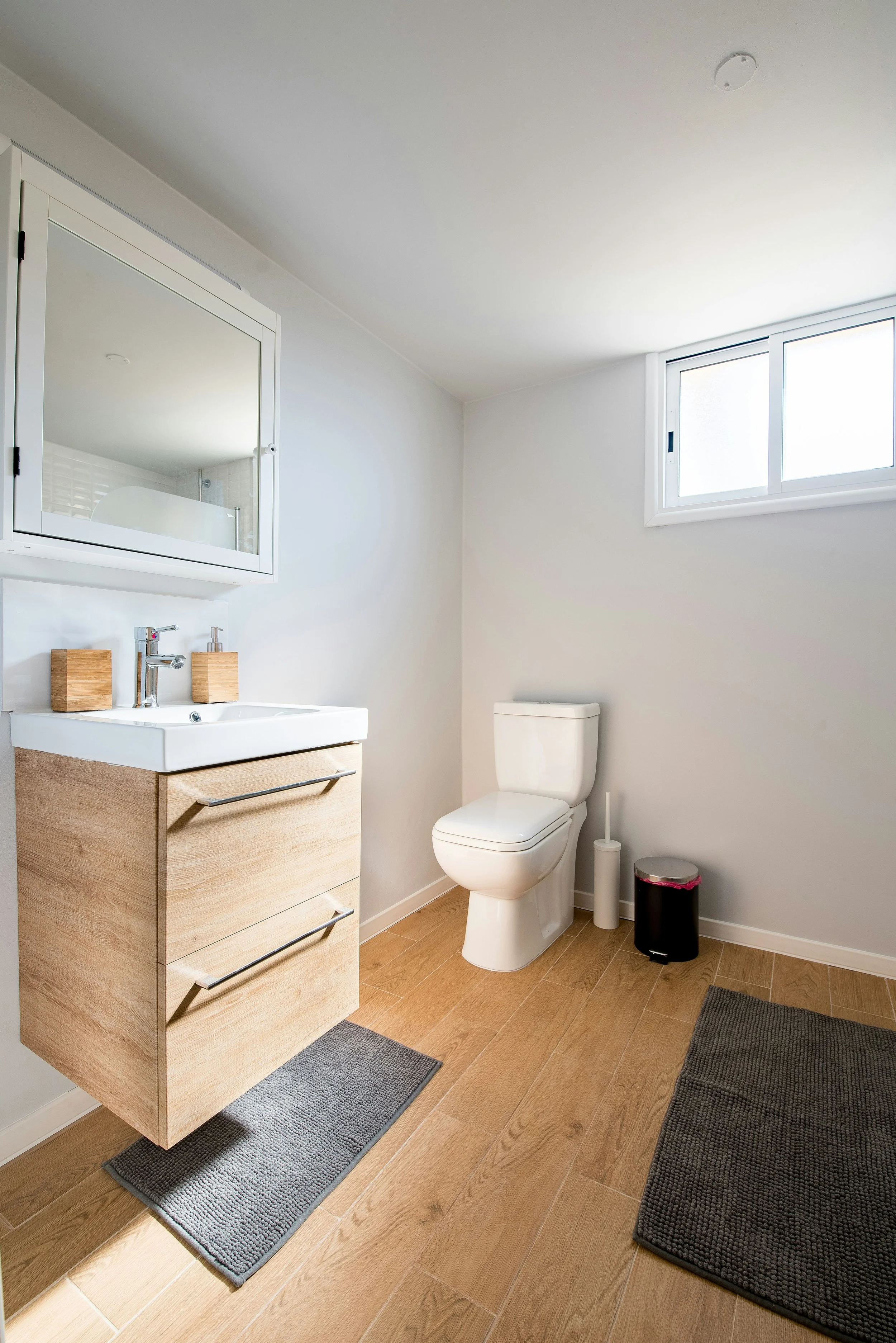 Modern bathroom with wooden fixtures, a white vanity, wall-mounted mirror, toilet, gray rugs, and a small window.