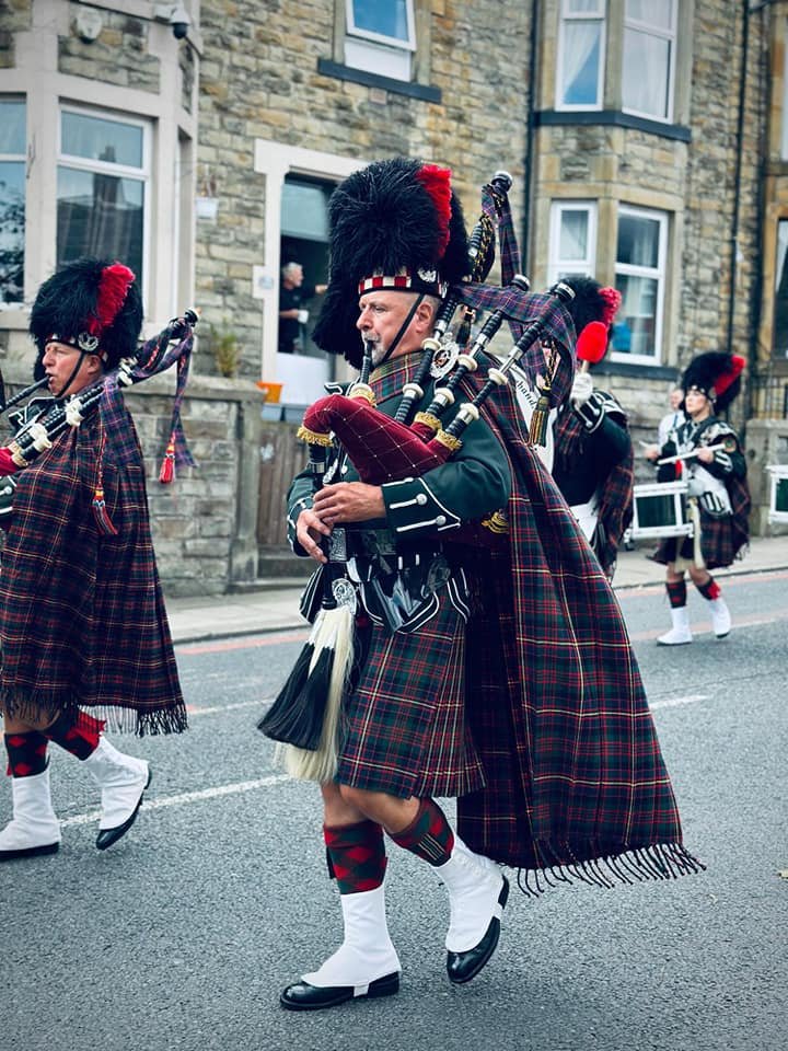A group of people dressed in traditional Scottish Highland dress, including kilts, socks, sporrans, and feathered hats, marching in a parade along a street with brick buildings in the background.