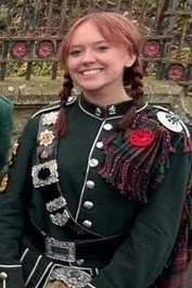 Young woman with red hair smiling, wearing a black Scottish military-style jacket decorated with medals and tartan shoulder sash, outdoors in front of a decorative fence.