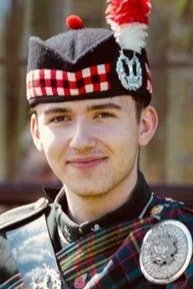 A young man in a military uniform wearing a glengarry hat with a black and red diced band, smiling outdoors.