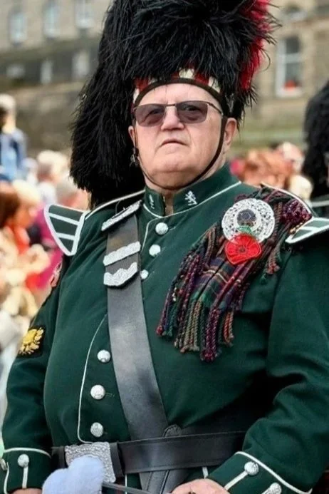 A woman dressed in a Scottish military uniform, including a green coat with silver buttons, shoulder epaulets, and a hat with a large black and red feather plume. She is wearing sunglasses and standing outdoors with a blurred crowd in the background.