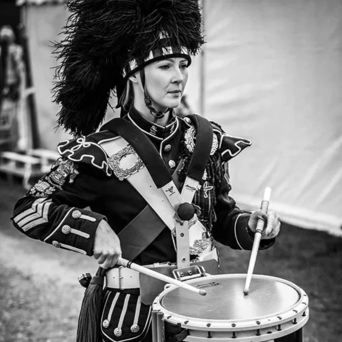Black and white photo of a woman in a historical military uniform playing a snare drum with drumsticks.