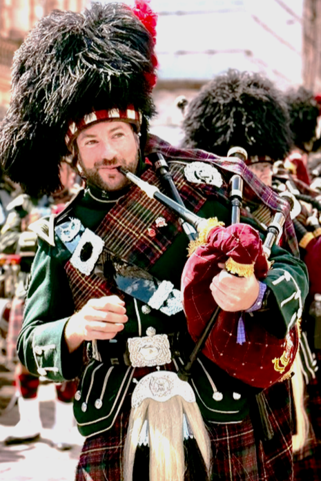 A man dressed in traditional Scottish Highland attire playing bagpipes, wearing a feather bonnet with red and white dicing, and a kilt with various accessories, during a parade or festival.