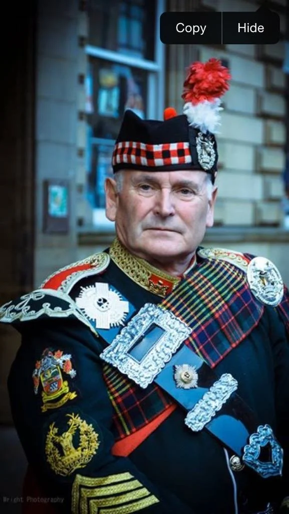 A man dressed in traditional Scottish military uniform with medals and patches, wearing a tartan sash and a Glengarry cap, standing outdoors in front of a building.