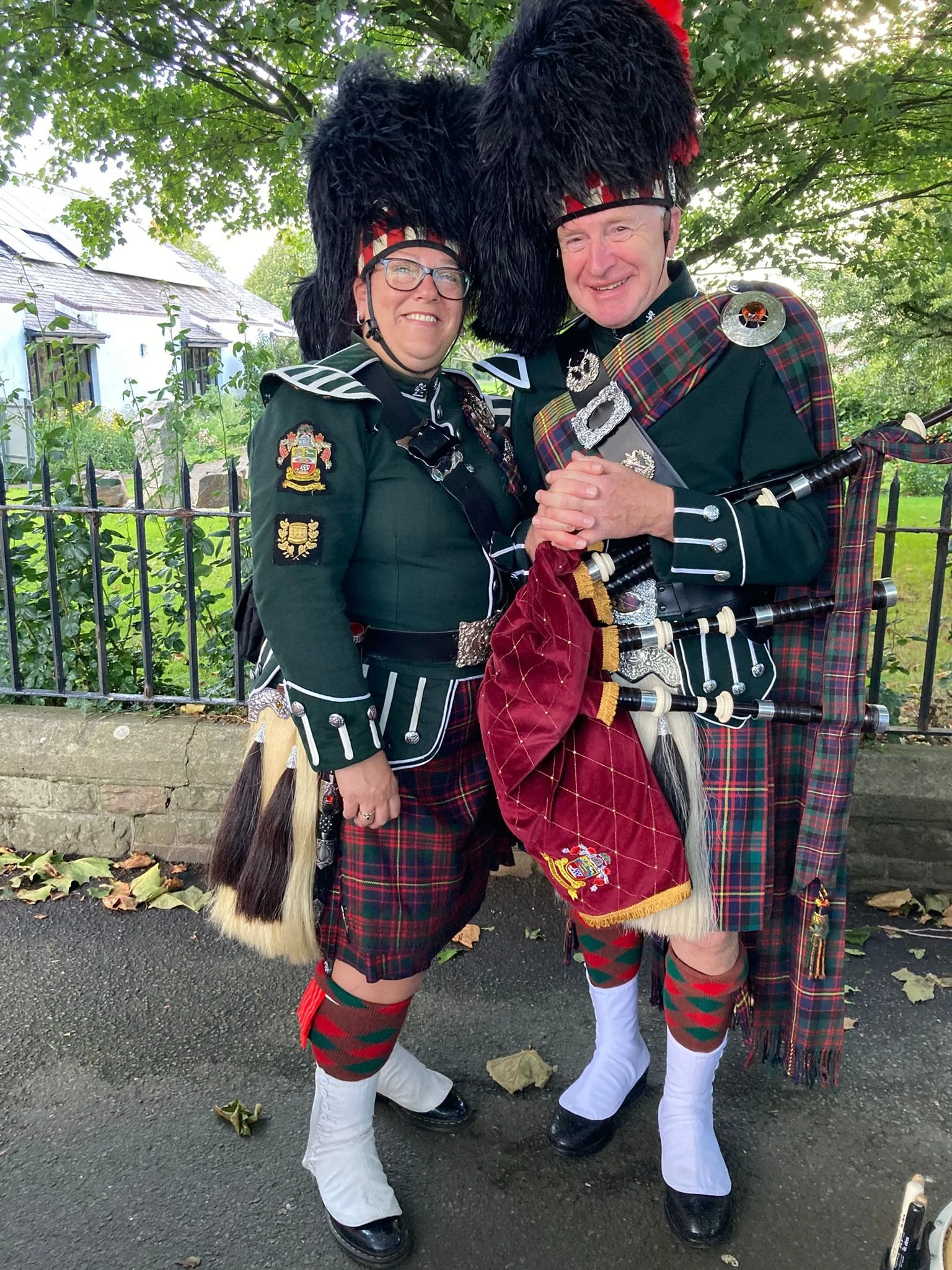 Two people dressed in traditional Scottish Highland attire, including kilts, sporrans, and feathered hats, standing outdoors with trees and a building in the background.