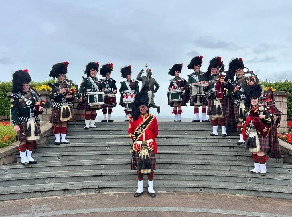 A group of people dressed in traditional Scottish Highland dress, including kilts, sporrans, and feathered hats, are standing on the steps of a monument or memorial, holding musical instruments.