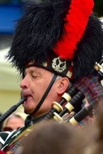 A man wearing a traditional Scottish uniform including a black feathered hat with red and black plumes playing the bagpipes.