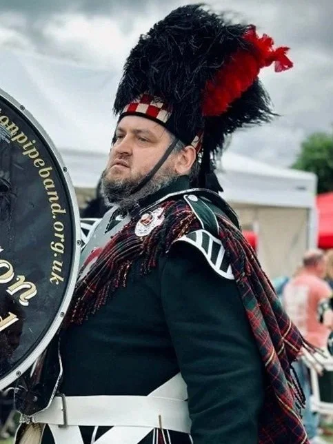 A man dressed in traditional Scottish military attire, including a black hat with red and black feathers, a tartan sash, and a dark uniform with white accents, standing outdoors with a cloudy sky and tents in the background.