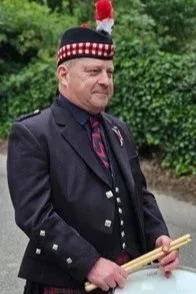 A man in traditional Scottish attire holding drumsticks, wearing a black jacket with silver buttons and a black and red checkered sash, outdoors with greenery in the background.