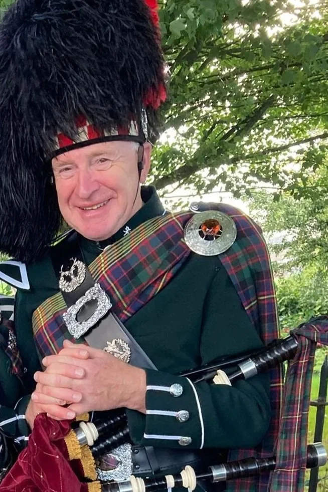 A man smiling in a Scottish military uniform, wearing a tall black and red feathered hat, a dark jacket with silver buttons, a tartan sash, and playing bagpipes outdoors.