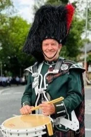 A soldier in a traditional Scottish Highland dress playing a drum outdoors, with trees and cars in the background.