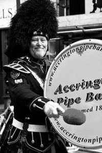 A man dressed in traditional Scottish Highland dress playing a large bass drum with the words 'Accrington Pipe Band' on it.