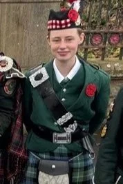 A young person in a green uniform with a red poppy and a square necktie, standing outdoors with a blurred background of a wooden fence and pink flowers.