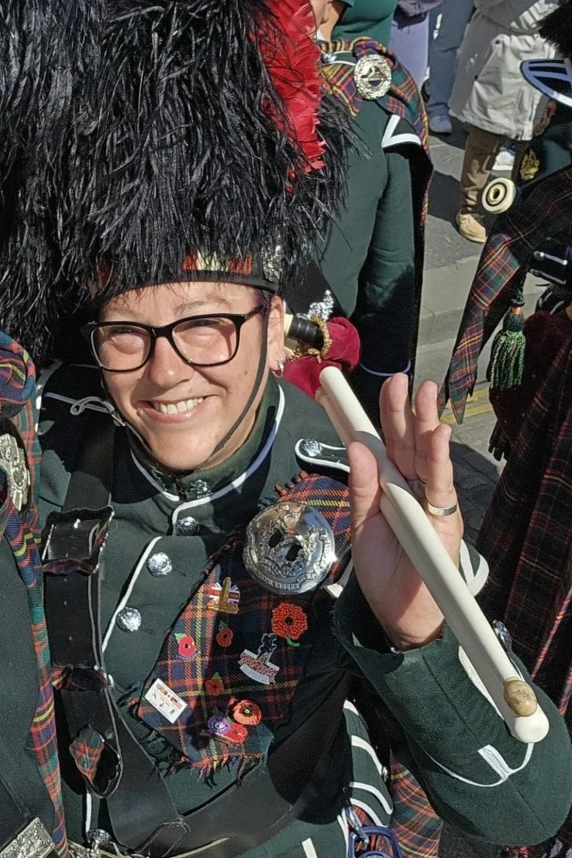 Person dressed in traditional Scottish attire, wearing a feathered hat, glasses, and a uniform with various badges, holding a ceremonial baton.