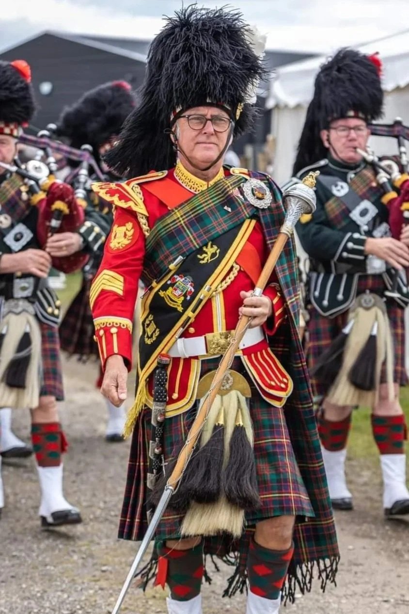 Man in traditional Scottish military uniform with a red jacket, tartan kilt, and badge, holding a staff, and wearing a tall black fur hat, participating in a Highland parade or ceremony.