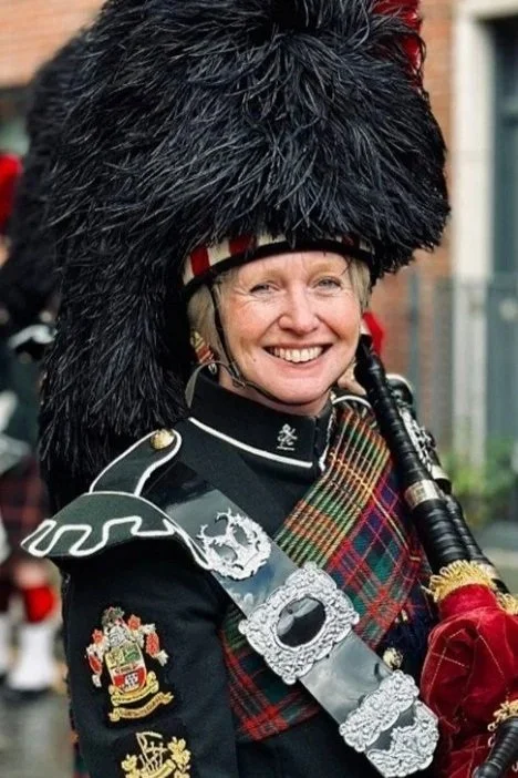 A woman dressed in traditional Scottish attire, smiling and holding bagpipes, wearing a tall feathered hat and a tartan sash.