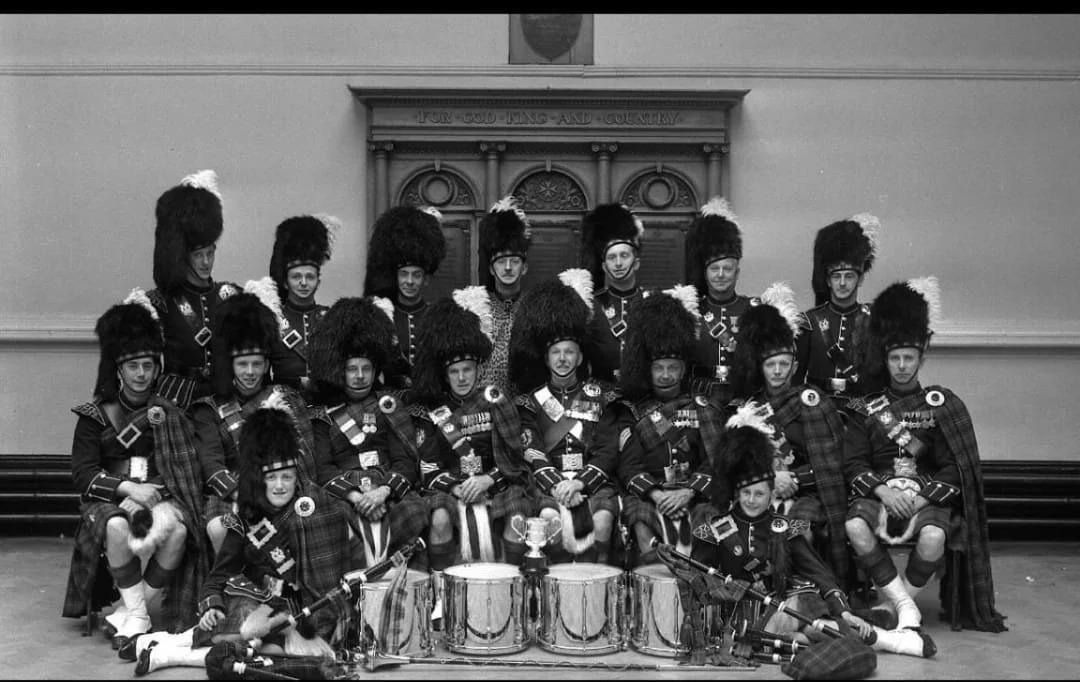 Group of boys in traditional Scottish Highland dress, including kilts and feathered hats, posing with drums in a hall.