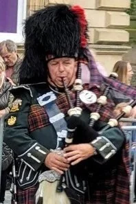 A person dressed in traditional Scottish Highland dress, including a feather bonnet and a tartan kilt, holding a bagpipe, at an outdoor event.