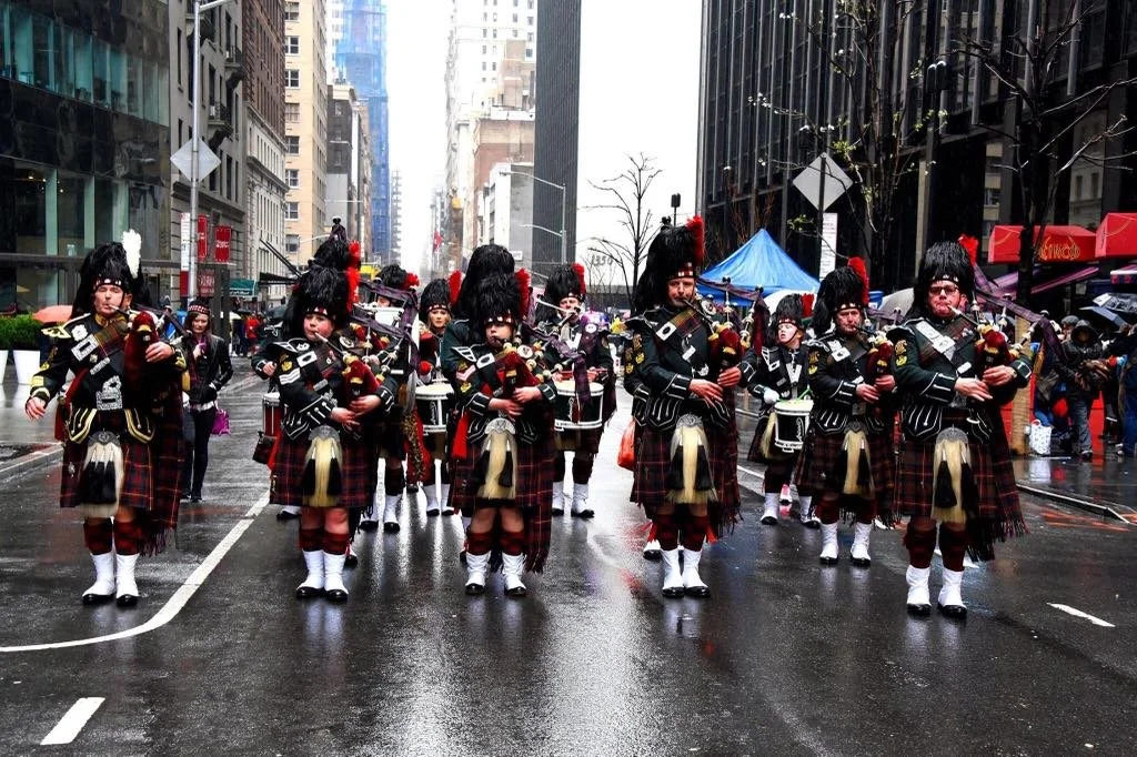 A marching band in traditional Scottish attire, including kilts and bearskin hats, parades down a city street on a rainy day.