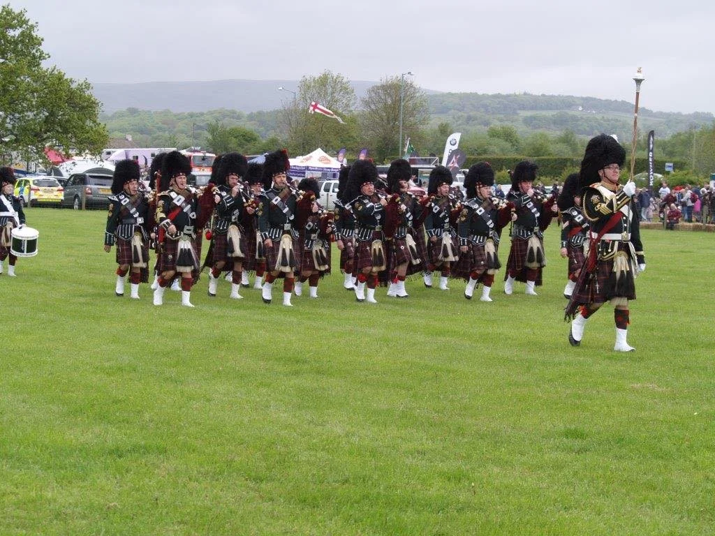 A group of people dressed in traditional Scottish Highland dress, including kilts and bearskin hats, marching across a grassy field during a parade or ceremony.