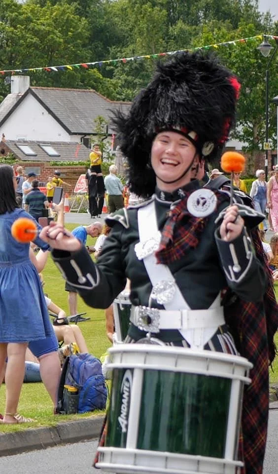 A person wearing a traditional Scottish military uniform, including a tall black fur hat and kilt, playing a snare drum at an outdoor event with people in the background.