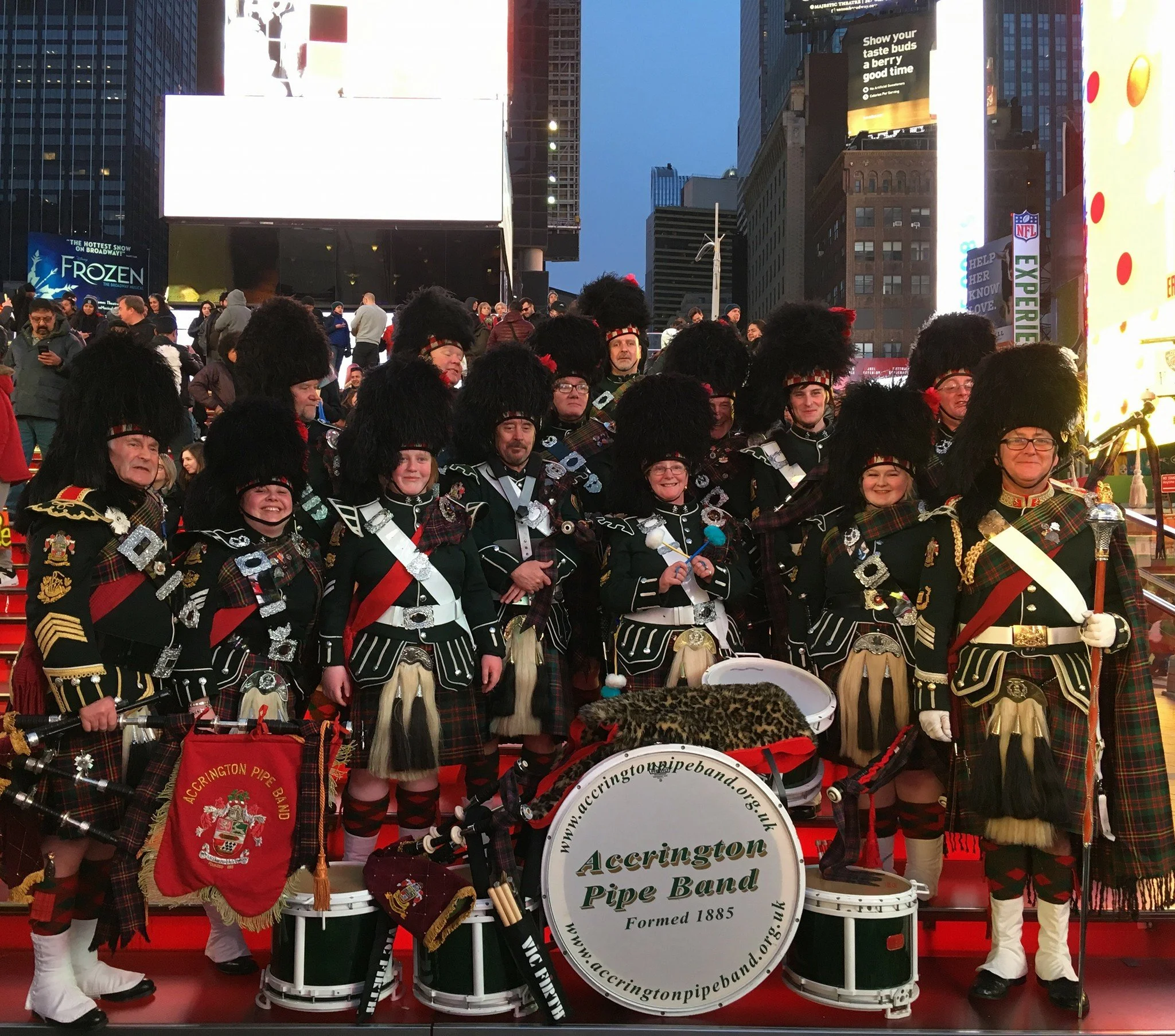 A group of people dressed in traditional Scottish attire, including kilts, sporrans, and tall fur hats, standing together on a red platform outdoors in an urban setting with tall buildings and electronic billboards in the background.