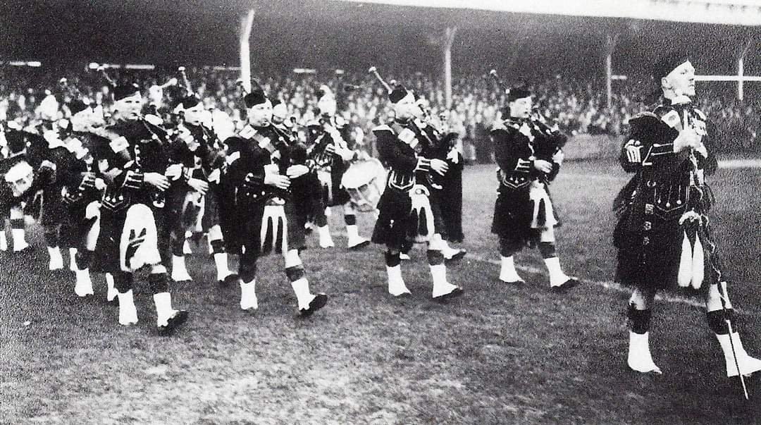 A black and white photograph of a marching band dressed in traditional Scottish attire, including kilts and sporrans, performing on a field with an audience in the stands in the background.