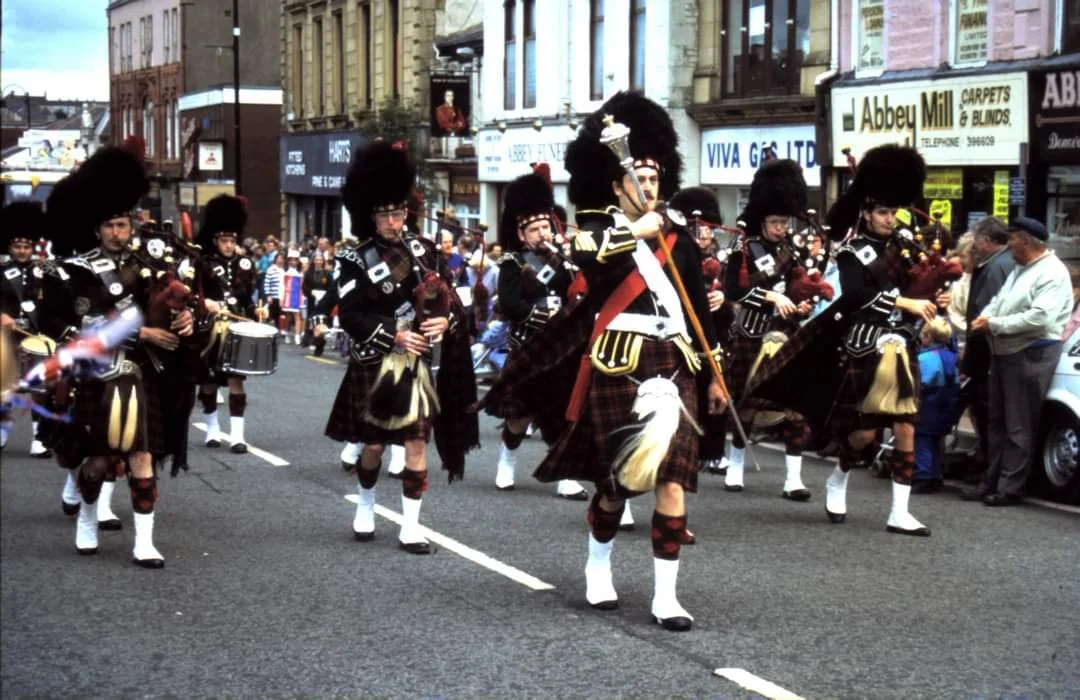 A parade featuring a group of people dressed in traditional Scottish attire, including kilts and tall hats, marching on a city street.