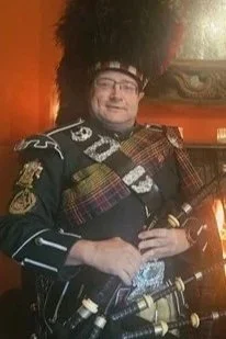 A person dressed in traditional Scottish attire, including a kilt and a sash, holding bagpipes, in front of a wooden-paneled wall.