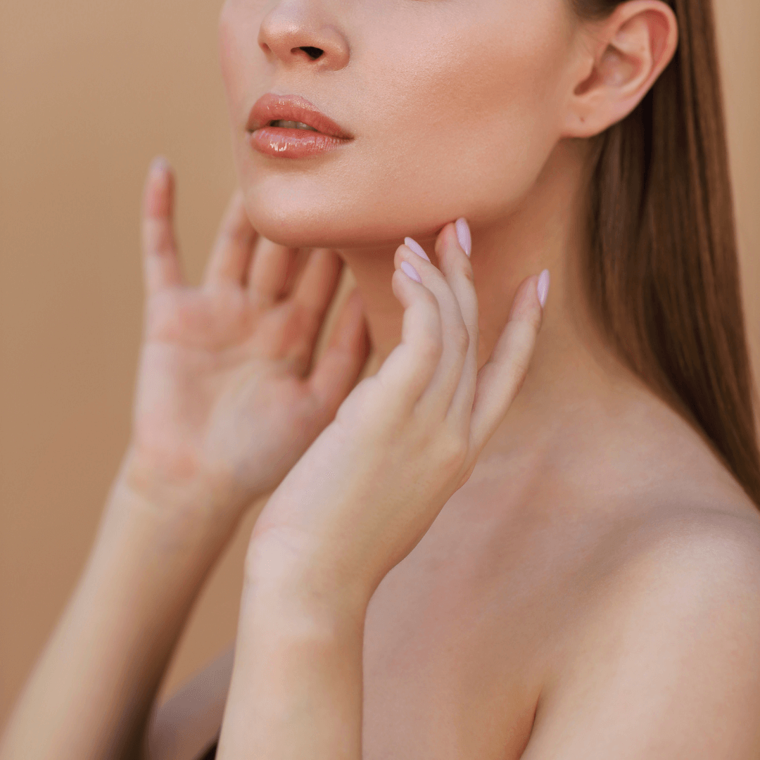 Close-up of a woman's lower face and neck with hands gently touching her jawline, showcasing smooth skin and soft natural makeup against a neutral background.