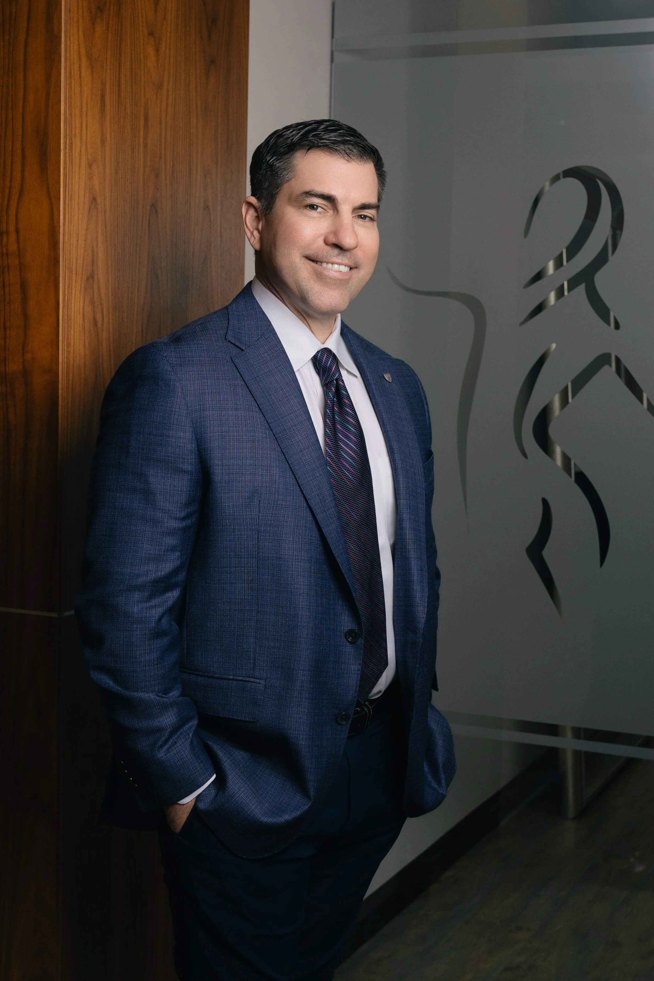 Man in blue suit standing indoors, smiling, with hands in pockets, wooden wall on left, frosted glass with design on right.