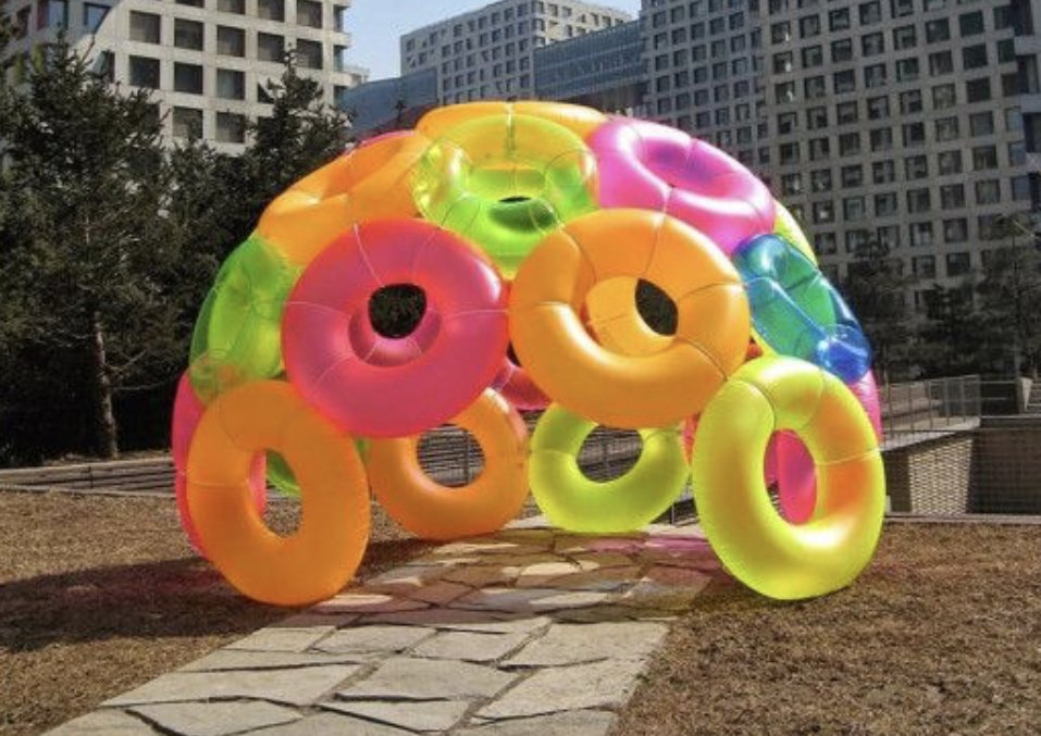 A colorful structure made of inflatable rings arranged in a dome shape outdoors, with buildings in the background.