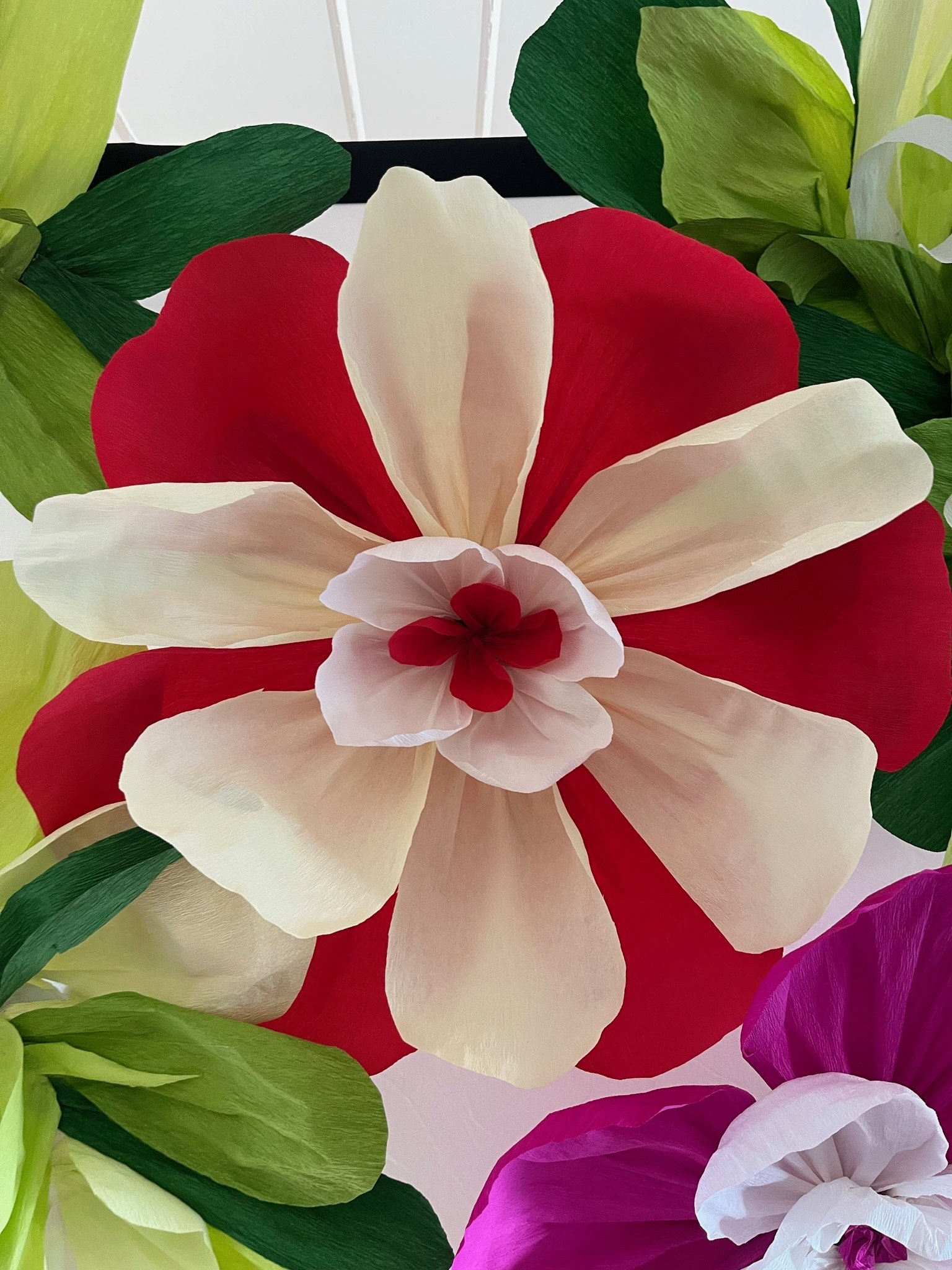Close-up of a large handmade paper flower with red and white petals, surrounded by green paper leaves and other colorful paper flowers.