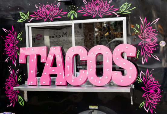 A taco truck window display with large pink letters spelling 'TACOS' surrounded by floral flower designs.