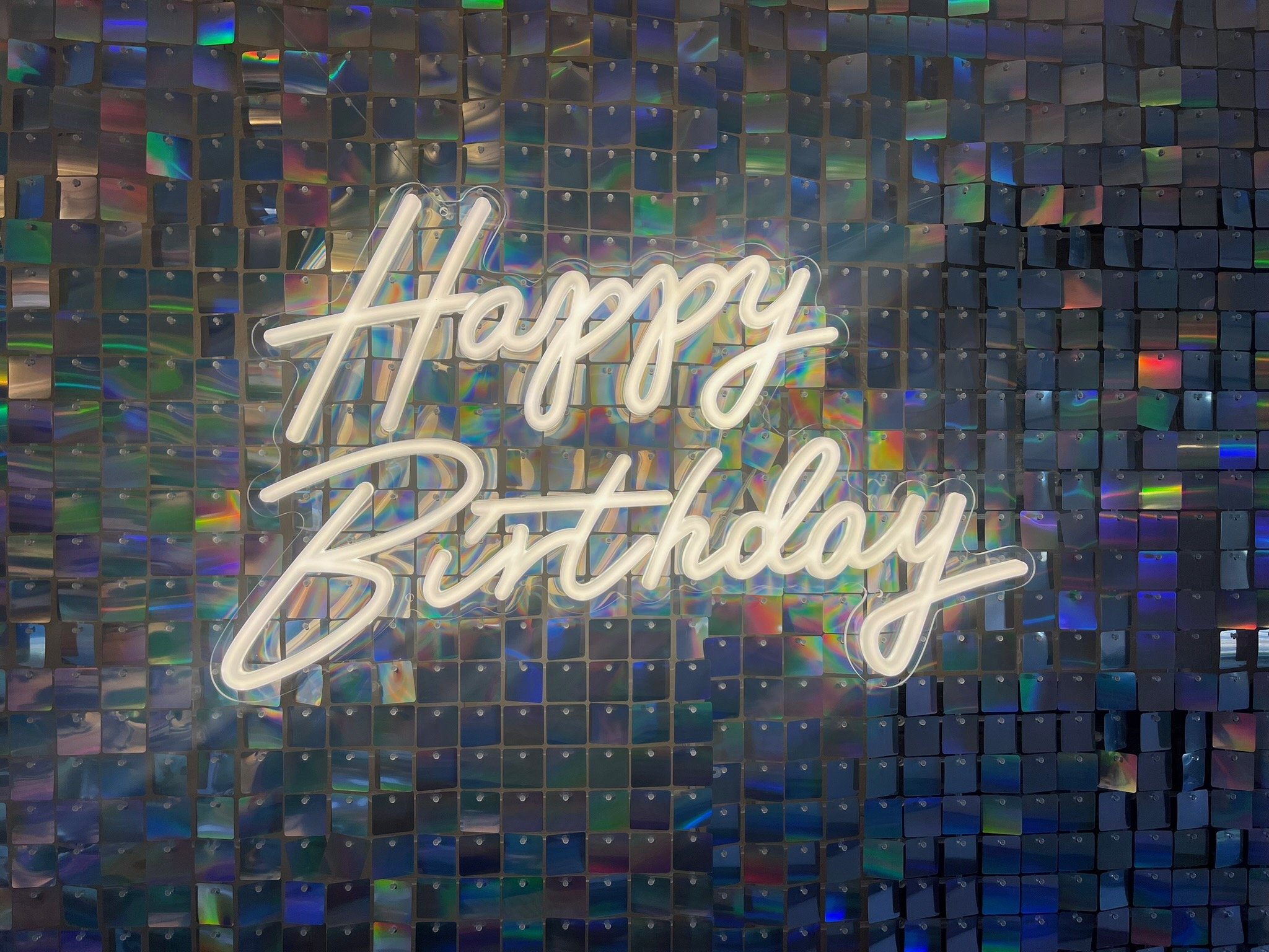 Neon sign with 'Happy Birthday' text on a holographic sequin backdrop.