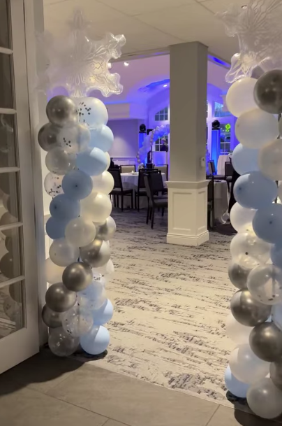 Decorated event entrance with balloon columns in white, blue, and silver colors, featuring large snowflake toppers, leading into a dimly lit room with tables and chairs.
