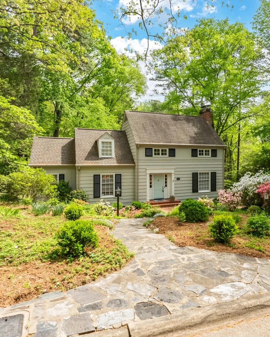 A professionally photographed two-story house in Chapel Hill, NC with beige siding, black shutters, and a brick step walkway leading to a white front door surrounded by a green garden, trees, and flowering bushes.