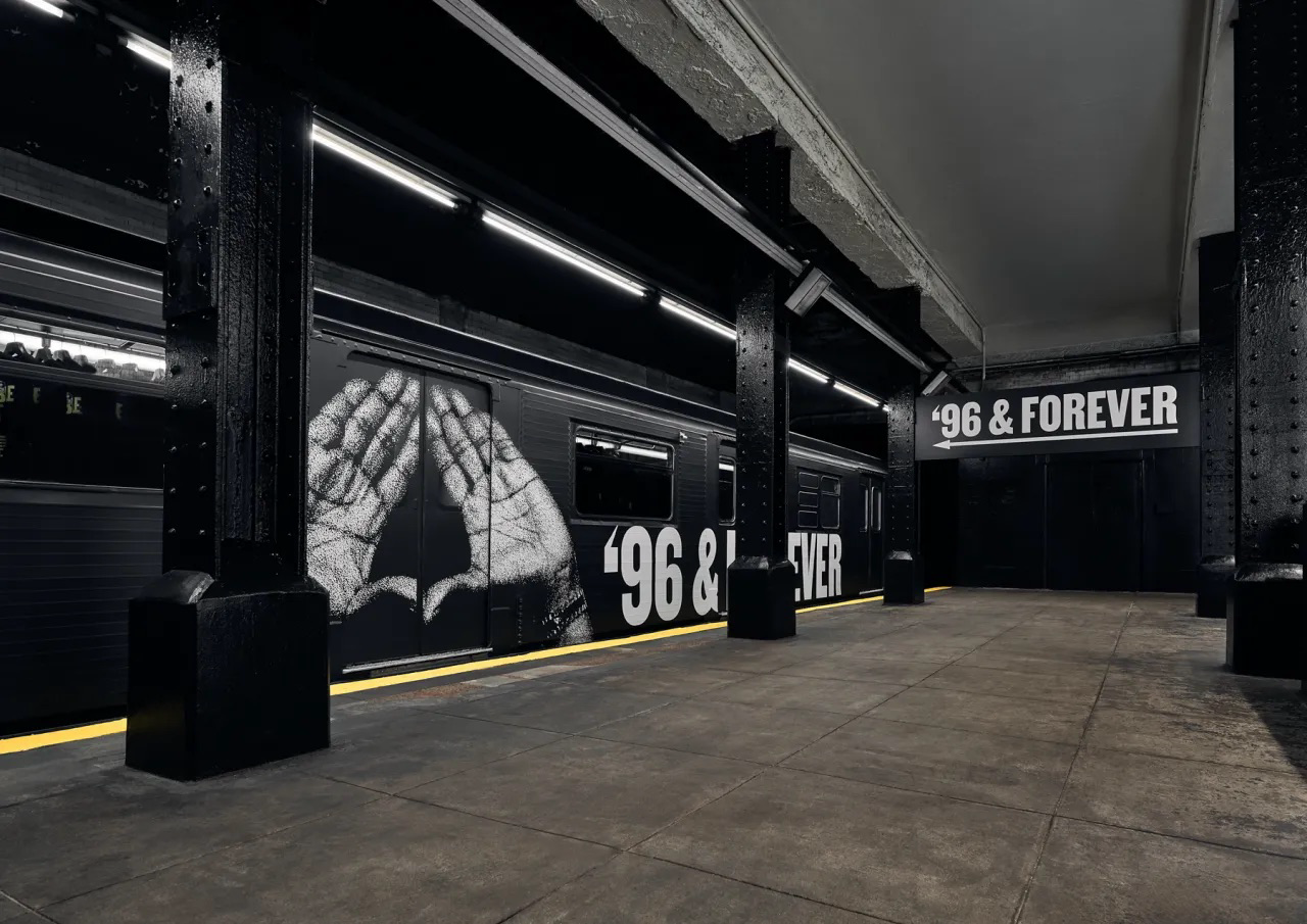 A subway station with black walls and pillars. Black and white mural of hands forming a triangle shape with the words '96 & FOREVER' on the wall and a sign overhead that reads '96 & FOREVER'.