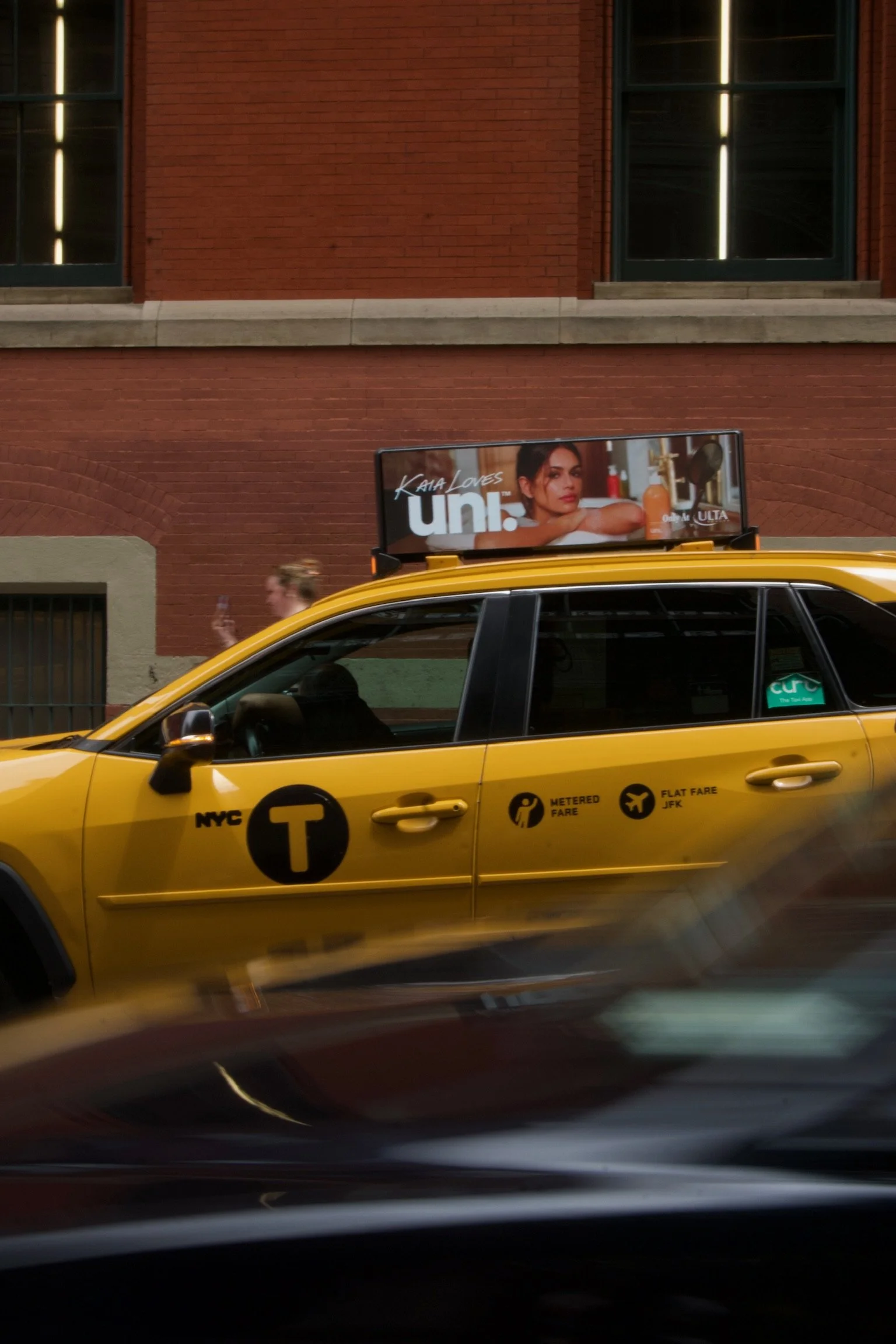 A yellow NYC taxi with a green sticker on the window, passing by a red brick building with large windows and a billboard on the roof featuring a Kaia Gerber with dark hair and a serious expression.
