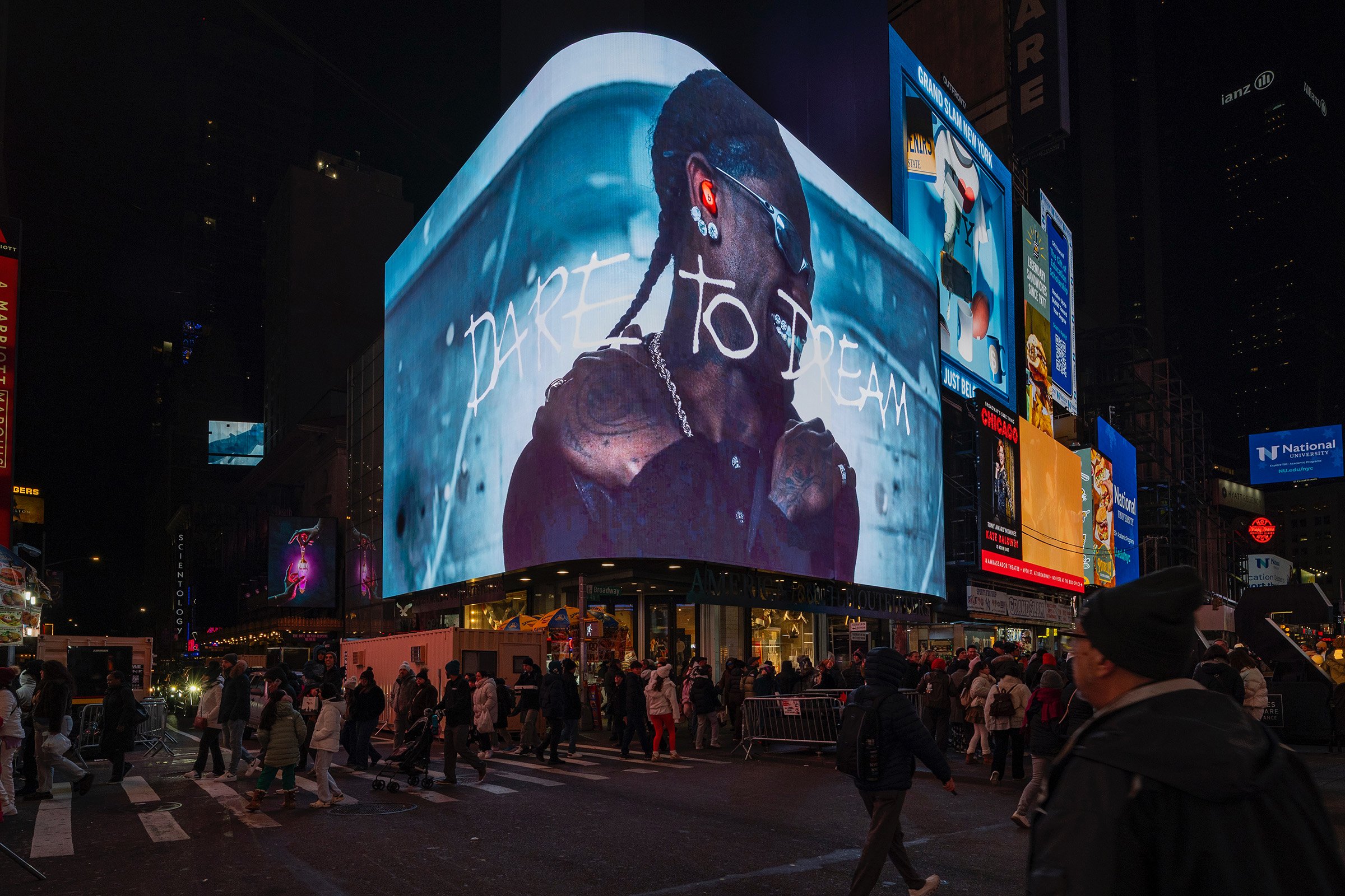 Night scene in Times Square with large digital billboard displaying Travis Scott with glasses, earrings, tattoos, and a silver chain, with the words 'Dare to Dream' written across the image. Crowds of people walk across the crosswalk below.