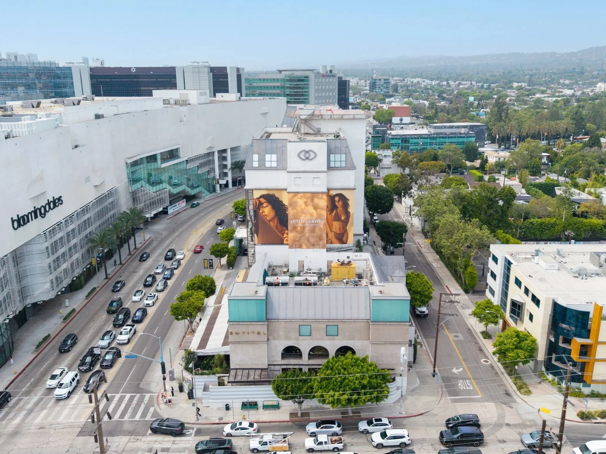 Cityscape featuring a large shopping mall with a prominent sign reading 'bloomingdales' on the left side, a busy street with many cars, and a building with an advertisement of a woman for Intimissimi. The background shows a sprawling city with trees 