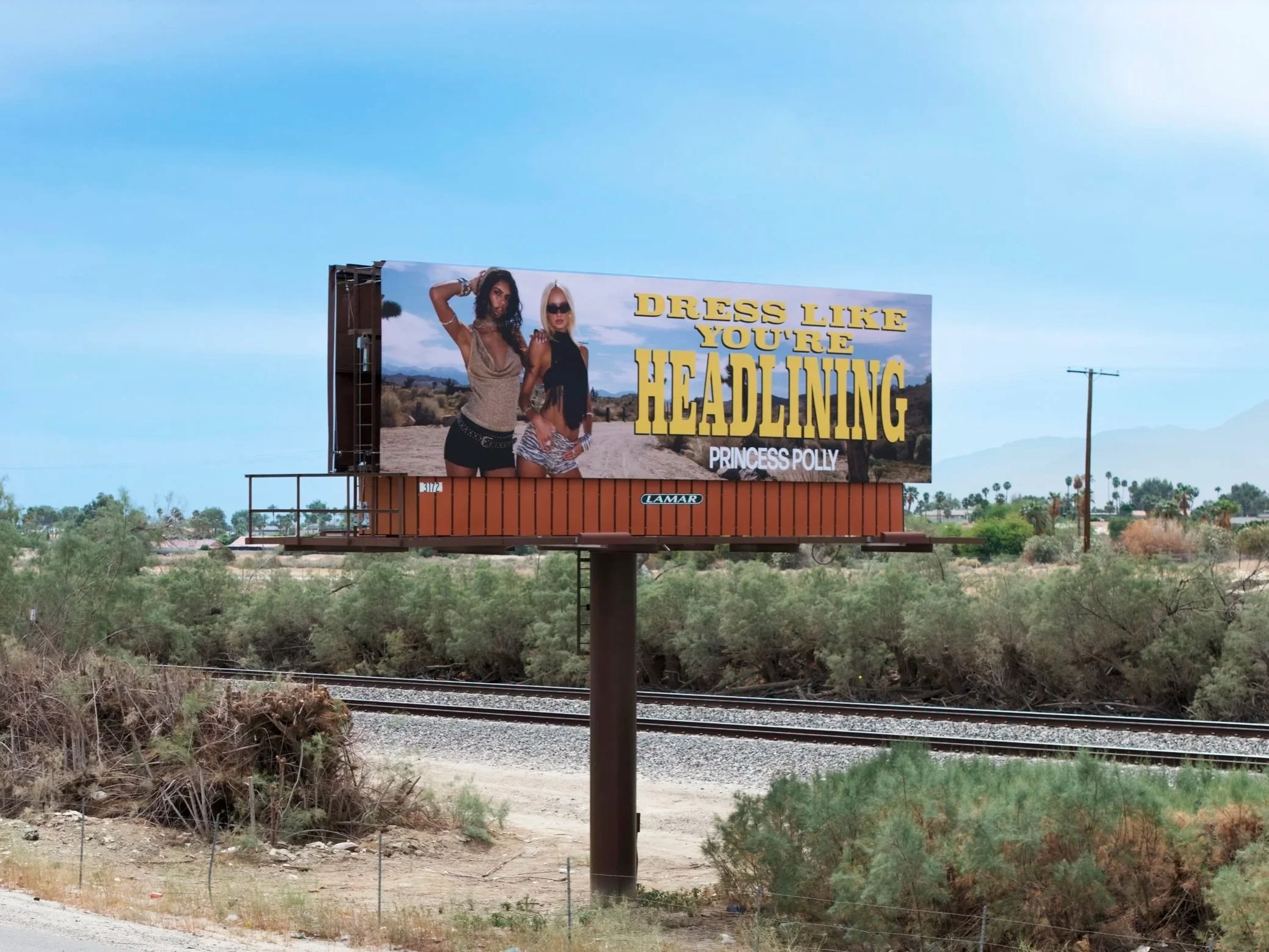 Billboard advertisement featuring two women, one with dark curly hair and the other with blonde straight hair, in a desert landscape with mountains in the background. The billboard has yellow text that reads, "DRESS LIKE YOU'RE HEADING" and smaller w