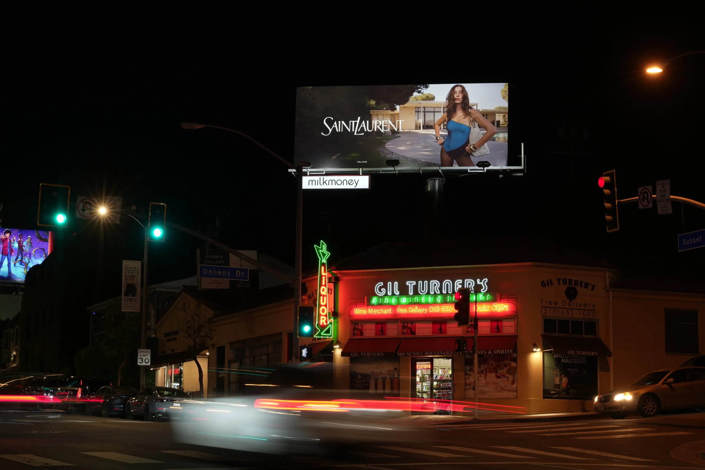Nighttime city street scene with traffic lights, and a restaurant with neon signs. There is a large billboard advertising Saint Laurent, featuring Hailey Bieber in a blue outfit. Vehicles and their light streaks are also visible.