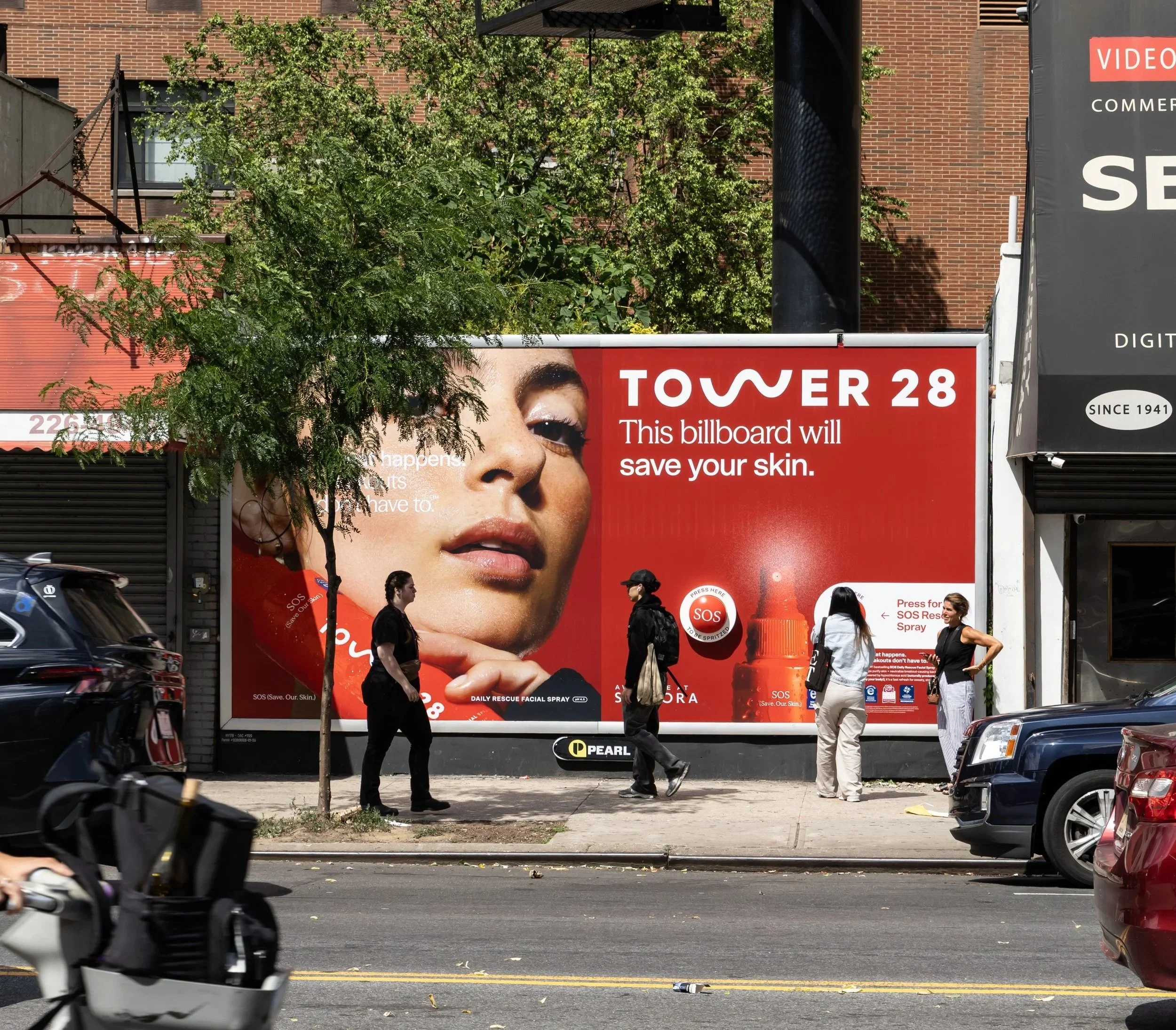 Street scene with a large interactive billboard for tower 28 and pedestrians walking, cars parked along the street, and a brick building in the background.