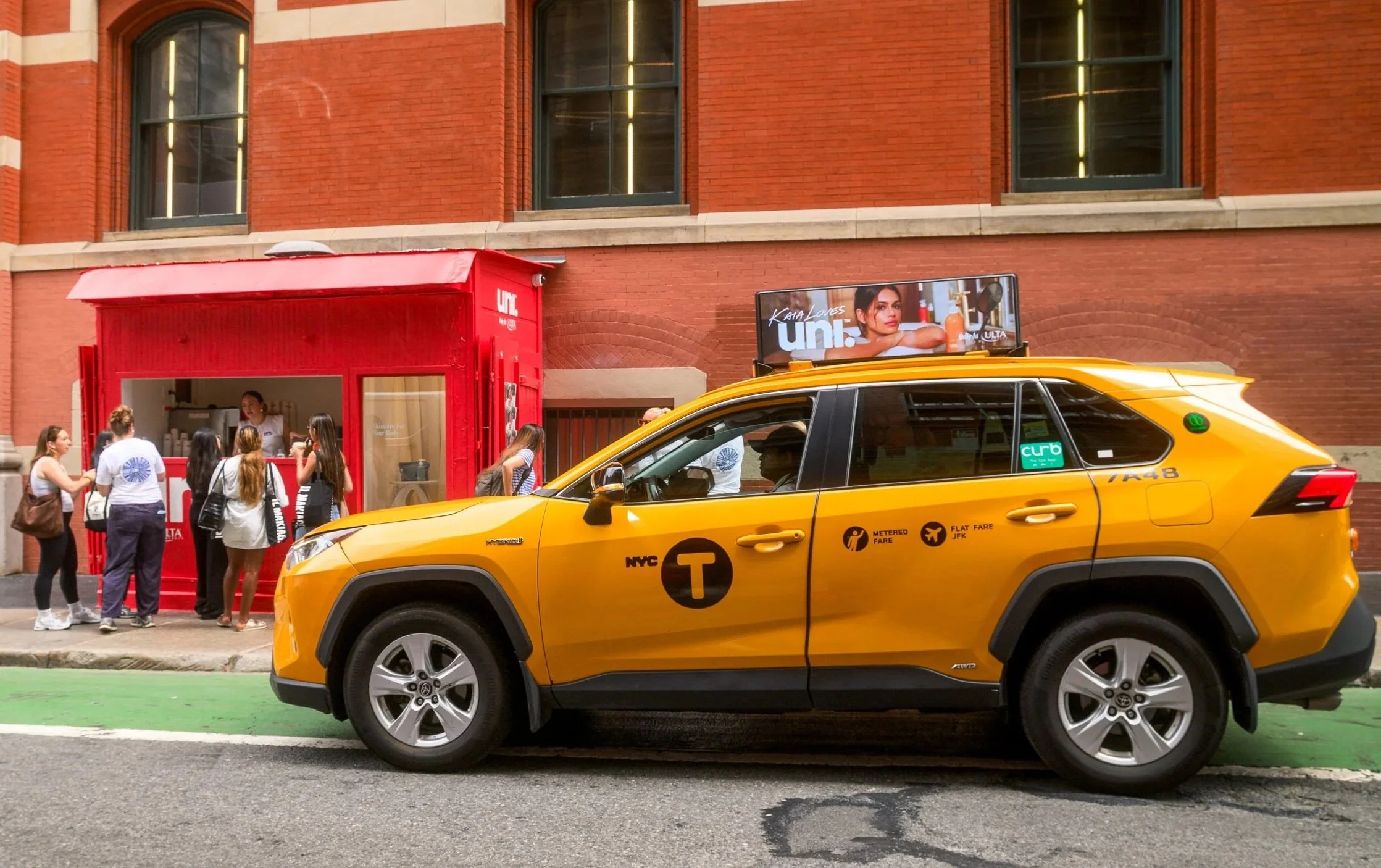 A yellow taxi cab driving on a city street with a group of people waiting in front of a red storefront and a billboard with Kaia Gerber's face on a brick building.