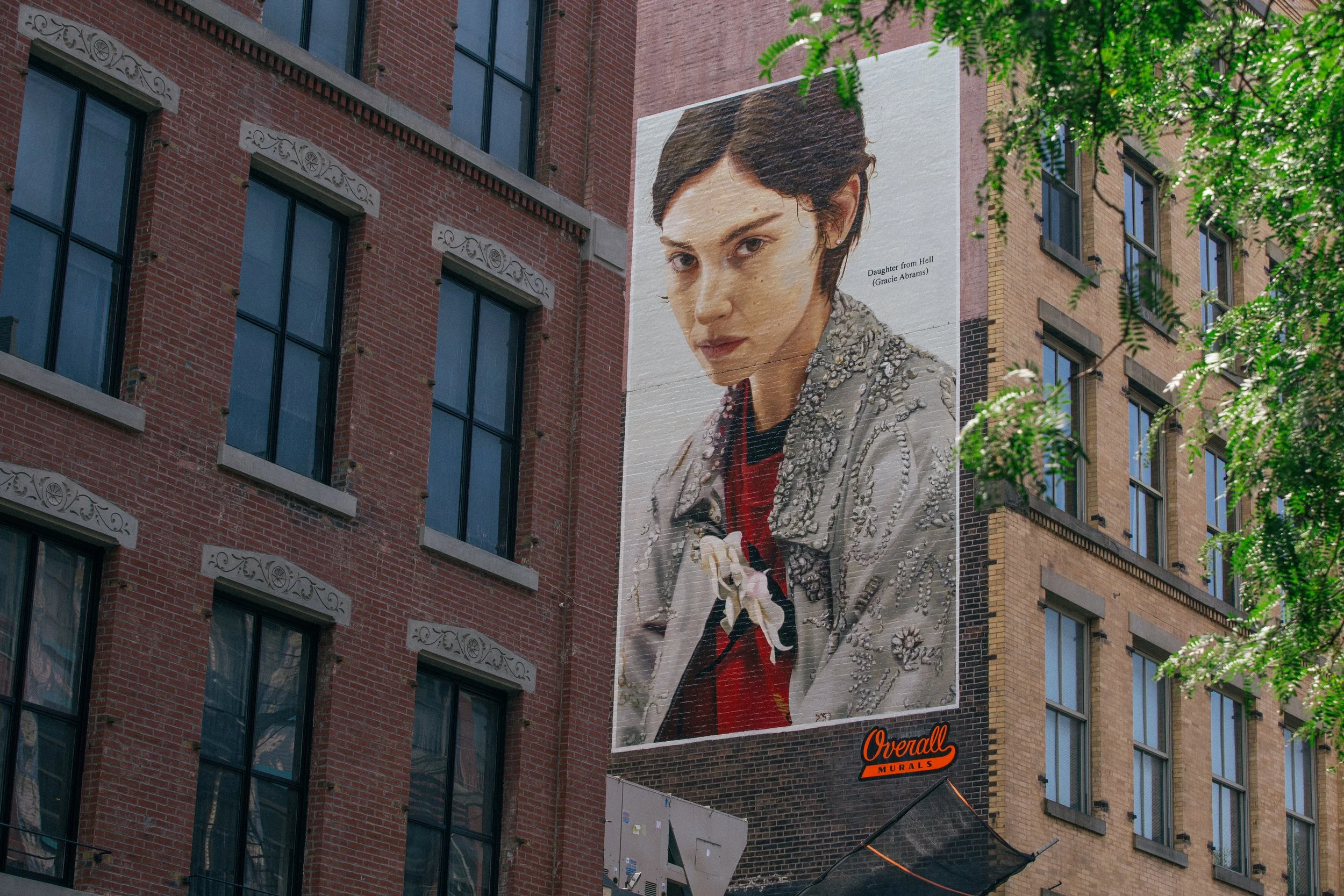 A large outdoor mural of a woman's face on the side of a brick building, with a quote from the movie 'Daughter from Hell' (Gracie Abrams). The mural depicts Gracie Abram's with short dark hair, wearing a shiny, beaded jacket.