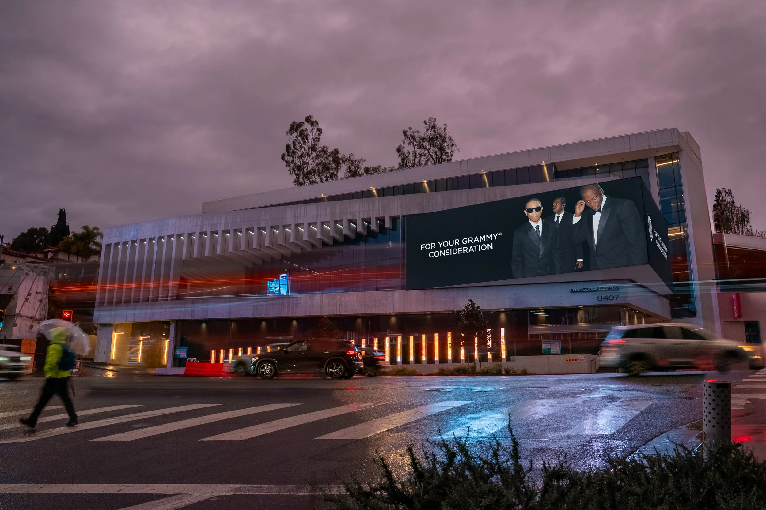 Nighttime city street scene showing a modern building with a large digital billboard featuring Pharrell Williams and text "For Your Grammy® Consideration". 