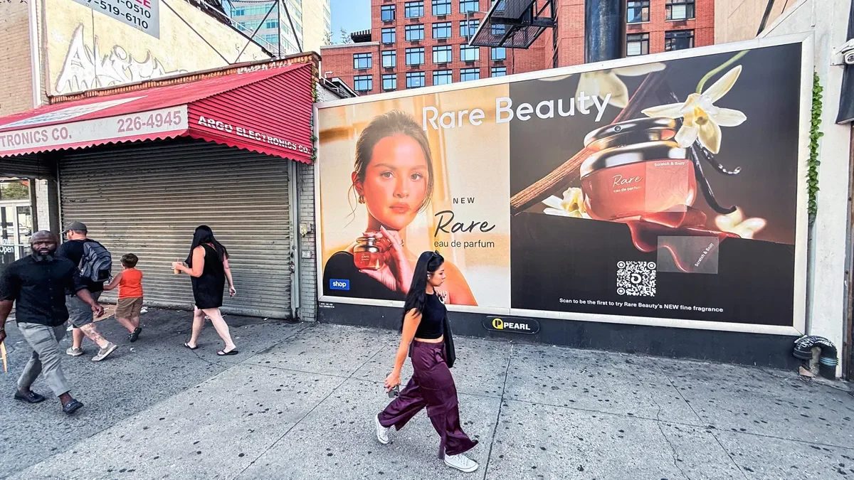A street scene with a large advertisement for Rare Beauty perfume on a wall with Selena Gomez.