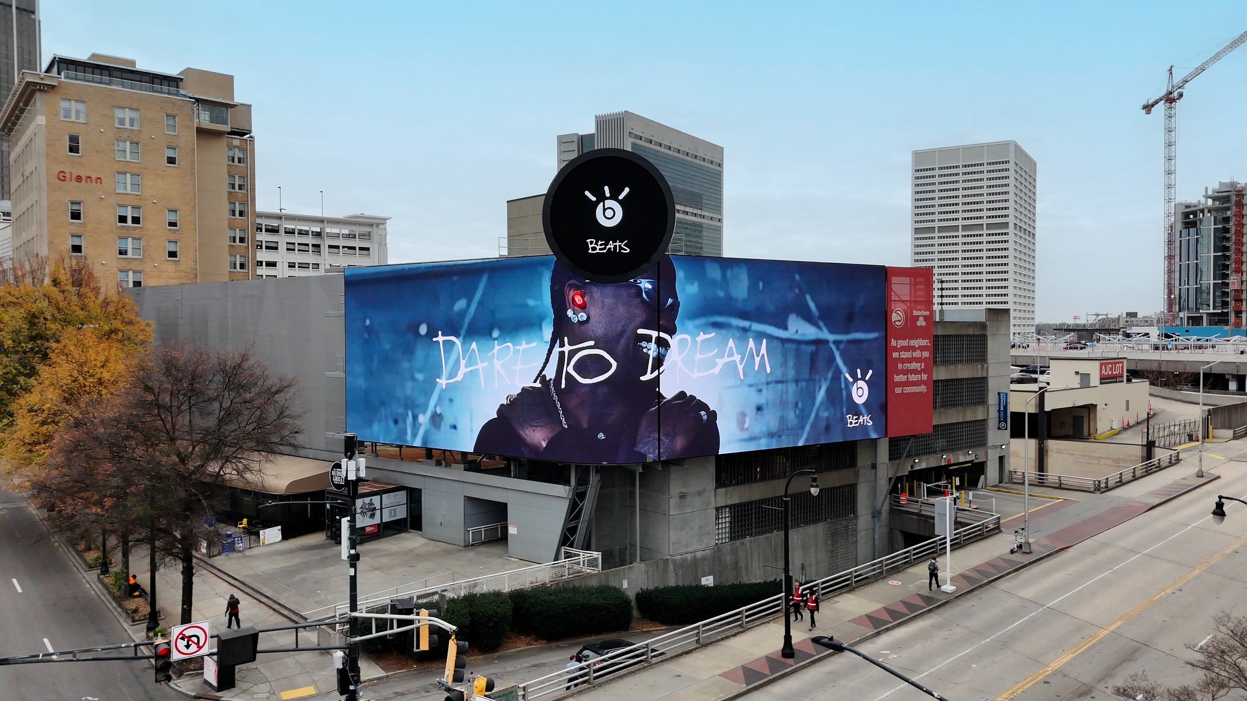 City street with a large digital billboard displaying an advertisement for Beats by Dre featuring Travis Scott wearing headphones. Surrounding buildings include a construction crane and a parking lot. Autumn trees line the sidewalk.