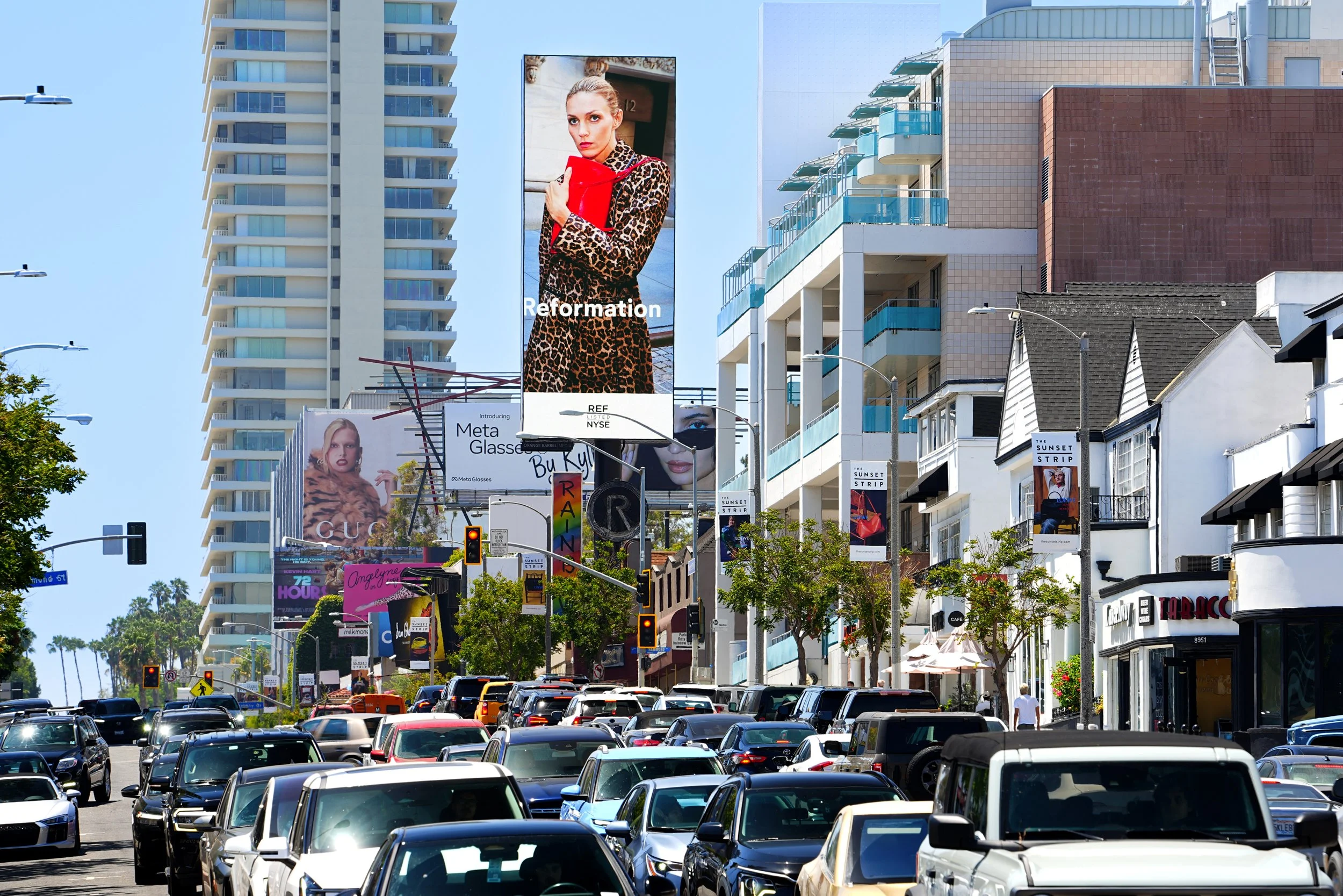 City street filled with cars and tall buildings. Large digital billboards display advertisements, including one with a woman in leopard print coat holding a red object with the word "Reformation."